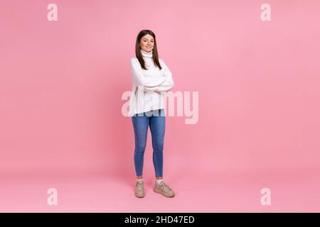 In voller Länge Porträt einer Frau, die mit gekreuzten Armen steht, Selbstvertrauen ausdrückt, die Kamera anschaut, einen weißen Pullover im lässigen Stil und eine Jeans trägt. Innenaufnahme des Studios isoliert auf rosa Hintergrund. Stockfoto