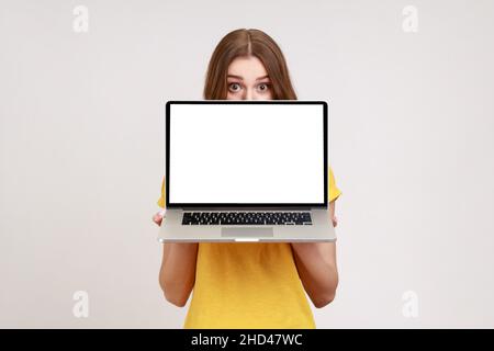 Positive unbekannte Frau in gelbem T-Shirt versteckt sich ein halbes Gesicht hinter dem Laptop mit weißem leeren Display, Blick auf die Kamera mit großen, überraschten Augen. Innenaufnahme des Studios isoliert auf grauem Hintergrund. Stockfoto