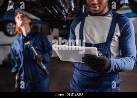 afroamerikanischer Mechaniker mit digitalem Tablet in der Nähe verschwommener Kollege und Auto in der Garage Stockfoto