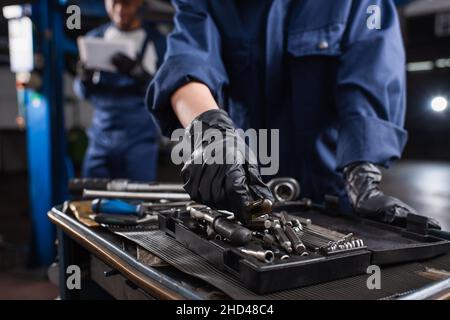 Beschnittene Ansicht des Mechanikers in Handschuhen, der das Werkzeug in der Nähe von verschwommenen Kollegen im Autodienst nimmt Stockfoto
