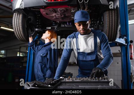 afroamerikanischer Mechaniker nimmt Werkzeug in der Nähe von Kollegen, die mit dem Auto in der Garage arbeiten Stockfoto