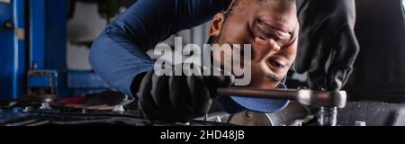 Positiver afroamerikanischer Arbeiter mit Schraubenschlüssel während der Arbeit im Autodienst, Banner Stockfoto