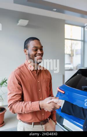 Zufriedener afroamerikanischer Mann schüttelt sich die Hände mit Autohändler in der Nähe verschwommenes Auto Stockfoto