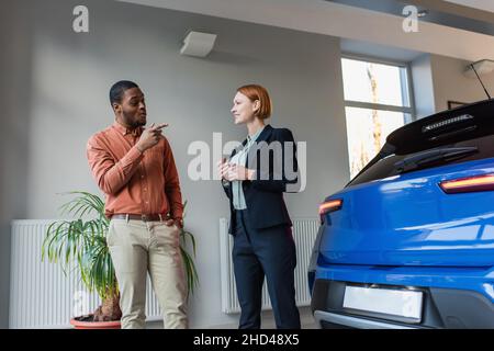 Erstaunlicher afroamerikanischer Mann, der mit dem Finger auf das Auto in der Nähe des lächelnden Autohändlers zeigt Stockfoto