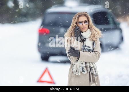 Unerfahrene weibliche Fahrerin, die das Auto von der rutschigen Winterstraße abstürzte, ruft den Abschleppdienst mit einem Notdreieck neben ihr vor dem Auto an. Stockfoto