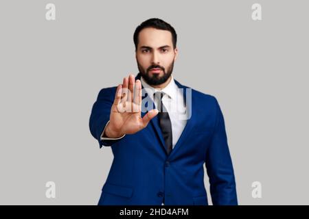 Ernsthafter bärtiger Mann, der eine Stop-Geste mit Handfläche macht, Konfliktverbotswarnung vor Gefahren, Mobbing aufhört und einen offiziellen Anzug trägt. Innenaufnahme des Studios isoliert auf grauem Hintergrund. Stockfoto