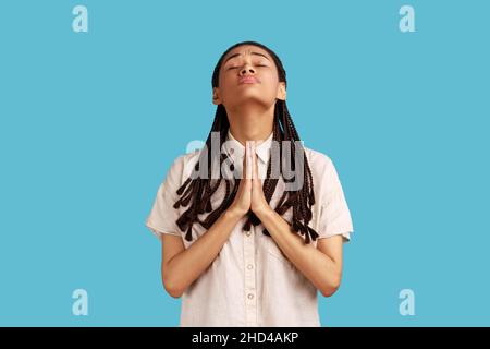 Frau mit schwarzen Dreadlocks, die die Hände im Gebet hält und fleht, mit hoffnungsvollen Augen aufschaut, sich entschuldigt, weißes Hemd trägt. Innenaufnahme des Studios isoliert auf blauem Hintergrund. Stockfoto