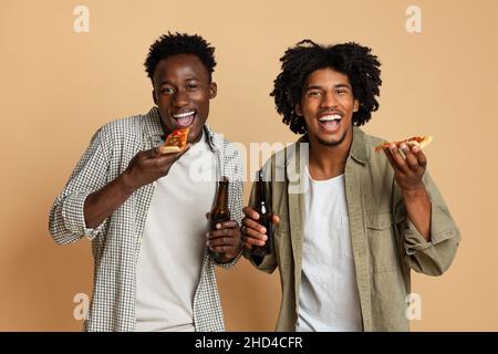 Freizeitkonzept für Herren. Zwei Schwarze Halten Bierflaschen Und Essen Pizza Stockfoto