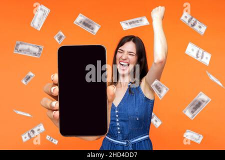 Online-Investitionen, Gewinner überglücklich reiche Frau und Geld verdienen. Porträt einer staunenden jungen Frau, die ein leeres mobiles Display zeigt und ihren Sieg feiert. Indoor isoliert auf orangefarbenem Hintergrund. Stockfoto