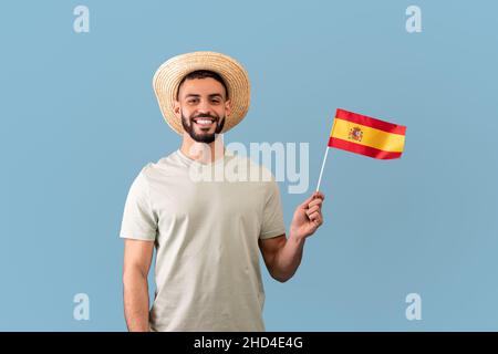 Glücklicher arabischer Mann in T-Shirt und Hut, hält die spanische Flagge und lächelt der Kamera zu, steht auf blauem Hintergrund Stockfoto