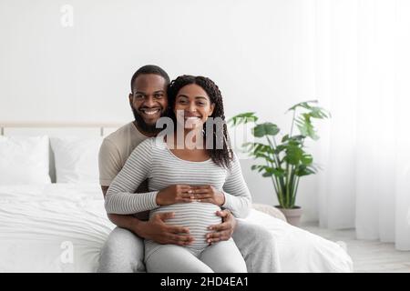 Zufrieden lächelnd gut aussehend Millennial african american Kerl umarmt schwanger Dame mit Bauch, sitzen auf dem Bett Stockfoto