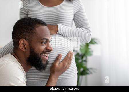 Frohes glückliches tausendjähriges afroamerikanisches Mann hört dem Bauch einer Schwangeren Frau im weißen Schlafzimmer, Nahaufnahme Stockfoto