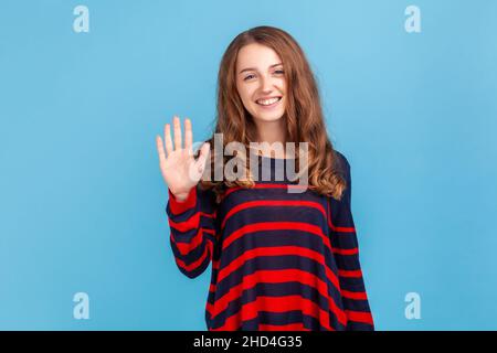 Porträt einer positiven jungen Frau, die einen gestreiften Pullover im lässigen Stil trägt, der mit winkender Hand steht und mit einem fesselnden, toothy Lächeln auf die Kamera blickt. Innenaufnahme des Studios isoliert auf blauem Hintergrund. Stockfoto