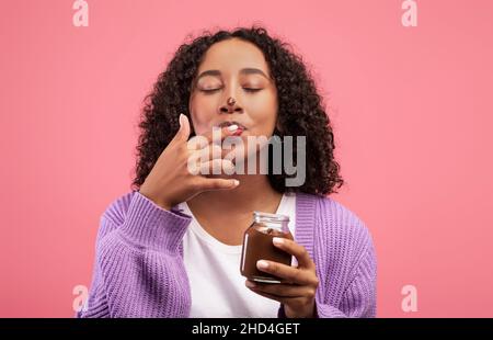 Schwarze Frau, die Schokoladenpaste aus dem Glas isst, Finger leckt, schmutzige Nase hat, Dessert auf rosa Hintergrund genießt Stockfoto
