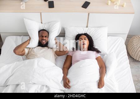 Wütend verärgerten tausendjährigen schwarzen Mann bedeckt seine Ohren mit Kissen, leidet unter Schnarchen und Lärm der Frau in der Nacht Stockfoto
