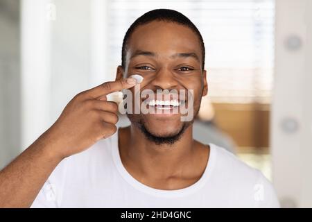 Nahaufnahme Eines Lächelnden, Hübschen Schwarzen Mannes, Der Feuchtigkeitscreme Auf Das Gesicht Auftragen Kann Stockfoto
