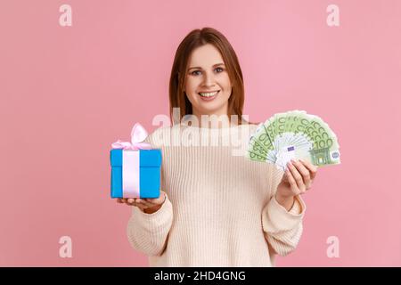 Porträt einer glücklichen optimistischen Frau, die eine eingepackte Schachtel und Euro-Banknoten hält, mit positivem Ausdruck die Kamera anschaut und einen weißen Pullover trägt. Innenaufnahme des Studios isoliert auf rosa Hintergrund. Stockfoto