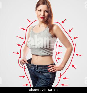 Porträt einer glücklichen, schönen, schlanken jungen Frau in großen Jeans und grauem Oberteil mit erfolgreicher Gewichtsabnahme, innen, Studioaufnahme, isoliert auf hellgrauem Hintergrund, Diät-Konzept. Stockfoto
