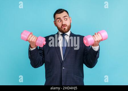 Porträt eines lustigen bärtigen Geschäftsmannes, der einen Anzug im offiziellen Stil trägt, in den Händen schwere rosa Hanteln hält und die Arme hebt, Training im Büro. Innenaufnahme des Studios isoliert auf blauem Hintergrund. Stockfoto