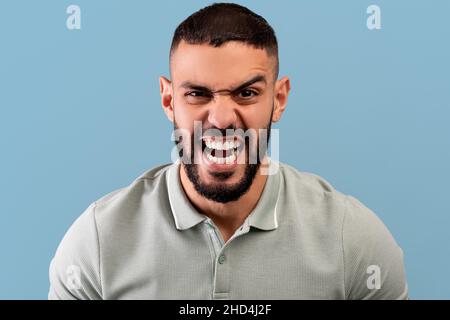 Porträt eines wütenden arabischen Mannes, der vor Wut auf die Kamera schreit, vor Wut schreit und auf blauem Hintergrund steht Stockfoto