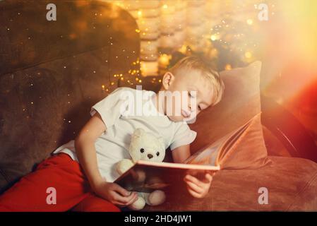 Der 3-4-jährige Junge liest ein Buch, das auf einem Sofa mit Teddybär sitzt. Home Lesung von Märchen. Netter kleiner Junge liest Buch, während er auf einem sitzt Stockfoto