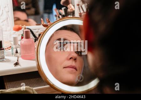 Porträt einer jungen Frau, die ihr Make-up vor einem runden, beleuchteten Spiegel mit LED-Beleuchtung macht Stockfoto