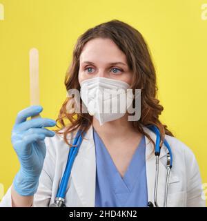 Eine Ärztin hält einen hölzernen medizinischen Spatel in der Hand. Medizinisches Instrument für die mündliche Untersuchung, gelber Studiohintergrund Stockfoto