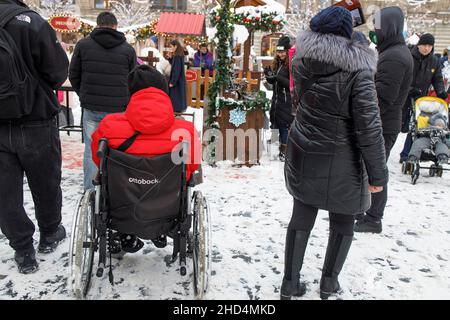 Moskau, Russland - am 20. Dezember 2021 Trägt Eine Frau einen Mann in einer roten Jacke mit einer Behinderung in einem Rollstuhl über den Roten Platz Stockfoto