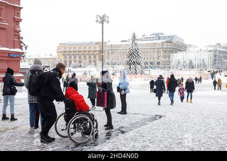 Moskau, Russland - am 20. Dezember 2021 Trägt Eine Frau einen Mann in einer roten Jacke mit einer Behinderung in einem Rollstuhl über den Roten Platz Stockfoto