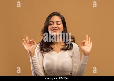 Meditationskonzept. Friedliche arabische Frau macht eine Geste und meditiert mit geschlossenen Augen vor beigem Hintergrund Stockfoto