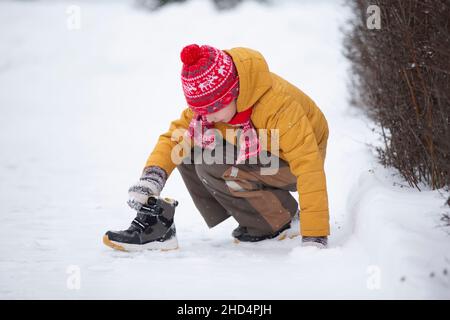 Ein kleiner Junge sitzt im Schnee und zieht einen Stiefel an. Kind im Winter. Stockfoto