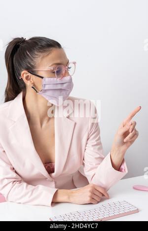 Salisbury, Wiltshire, Großbritannien. 2021. Attraktive Frau trägt rosa Gesichtsmask und zeigt auf Computer und Tastatur. Stockfoto