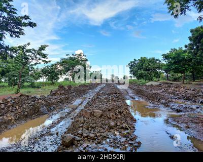 Stock Foto von chaotisch schlammigen Feldweg nach starkem Regen mit tiefen Reifen Spuren in der Landschaft, Straße von grünen Bäumen und Ackerland umgeben . blu Stockfoto