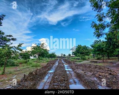 Stock Foto von chaotisch schlammigen Feldweg nach starkem Regen mit tiefen Reifen Spuren in der Landschaft, Straße von grünen Bäumen und Ackerland umgeben . blu Stockfoto