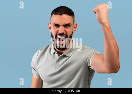 Wütender Mann aus dem Nahen Osten schüttelt geballte Faust auf die Kamera und schreit und zeigt seinen Protest auf blauem Hintergrund Stockfoto