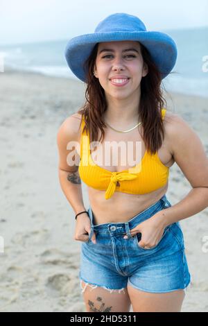 Porträt einer jungen Frau am Strand in blauem Hut, Shorts und Badeanzug Stockfoto