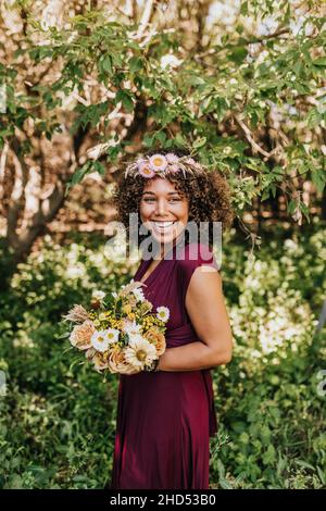 Lächelnde Frau steht im Wald mit Blumenstrauß und trägt die Blumenkrone Stockfoto