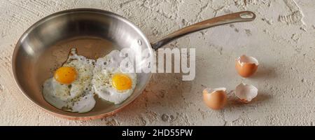 Zwei Spiegeleier in Kupferpfanne und Eierschalen auf rauem, texturiertem Tongrund. Gesundes Frühstückskonzept. Stockfoto