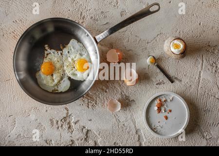 Gebratene zwei Eier in Kupferpfanne und hartgekochtes Ei in Steinzeug-Eierbecher auf rauem, texturiertem Lehm-Hintergrund. Gesundes Frühstückskonzept. Stockfoto