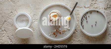 Ein gekochtes Ei in Steinzeug Eierbecher mit Eierschale zerbrochene Stücke in Betonschale, Salzflocken und Pfeffer in weißen Betonschüsseln auf grob strukturiertem cl Stockfoto