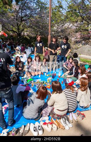 Eine Gruppe japanischer Teenager auf einer Kirschblütenparty, die auf einem Quadrat auf einer blauen Matte sitzt, während einige andere in der Frühlingssonne herumstehen. Stockfoto