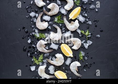 Meeresfrüchte Hintergrund für die Präsentation Ihrer Produkte. Gefrorene Ozeanjumbo-Garnelen mit Eis und Zitrone auf schwarzem Hintergrund zum Kochen. Pazifische weiße Garnelen Stockfoto