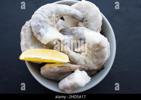 Gefrorene große Garnelen in einer grauen Schüssel auf schwarzem Hintergrund mit Eis, Zitrone zum Kochen. Köstliche Königsgarnele ohne Kopf für die Zubereitung von Mahlzeiten. Genießen Sie die natürliche Natur Stockfoto