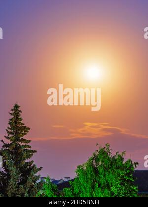 Mond und diffuses Mondlicht in einem violetten Himmel unter den Dächern von Häusern und Bäumen Stockfoto