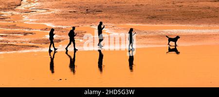 Ein Panoramabild einer Familie von Urlaubern, die am Fistral Beach in Cornwall spazieren gehen und von einem intensiven Sonnenuntergang umgeben sind. Stockfoto