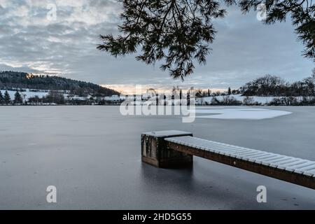 Hölzerner Pier am See mit frischem Schnee.Winterteich mit kleinem Steg bei Sonnenaufgang, Dorf im Hintergrund.Frostige ruhige Landschaft. Weiße Winterlandschaft verschneit Stockfoto