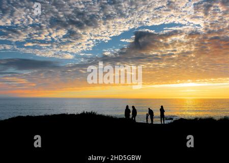 Urlauber, die an der Küste stehen, werden von einem wunderschönen, farbenfrohen Sonnenuntergang über Little Fistral an der Küste von Newquay in Cornwall verzaubern. Stockfoto
