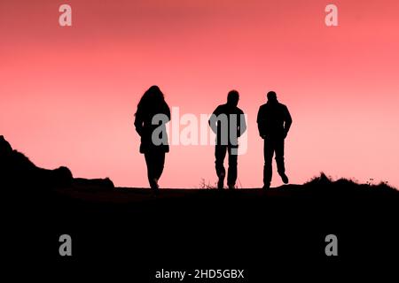 Drei Menschen, die am Ende des Tages in Newquay in Cornwall den Küstenpfad entlang wanderten, schillerten gegen einen bunten Himmel. Stockfoto