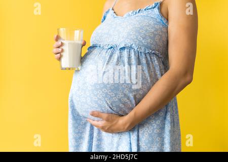 Croped Portrait niedlich schwanger unkenntlich Frau in floralen blauen Kleid halten trinken Glas Milch auf gelbem Hintergrund kopieren Raum. Mutterschaft Stockfoto