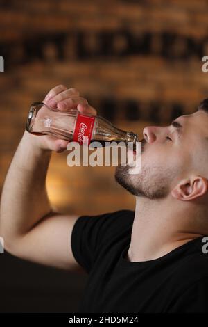 RIVNE, UKRAINE JUNI 26 2020: Junger bärtiger Mann trinkt Coca Cola aus der Flasche im Friseurladen auf Backsteinmauer Hintergrund. Stockfoto